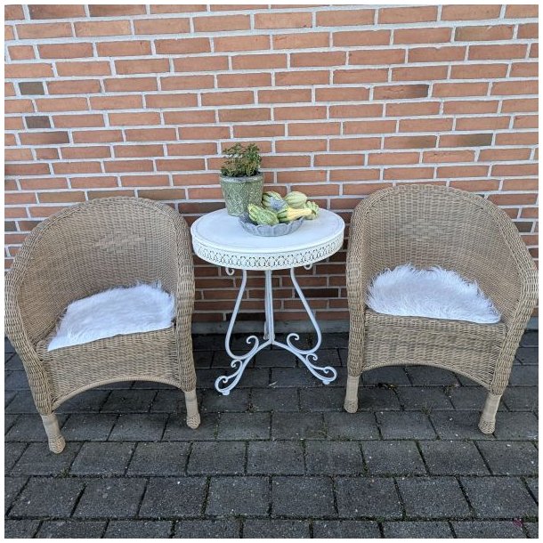 To naturfarvede flettede havestole placeret ved et bord, hvor den forreste stol er stylet med et firkantet hvidt imiteret lammeskind som s�dehynde.