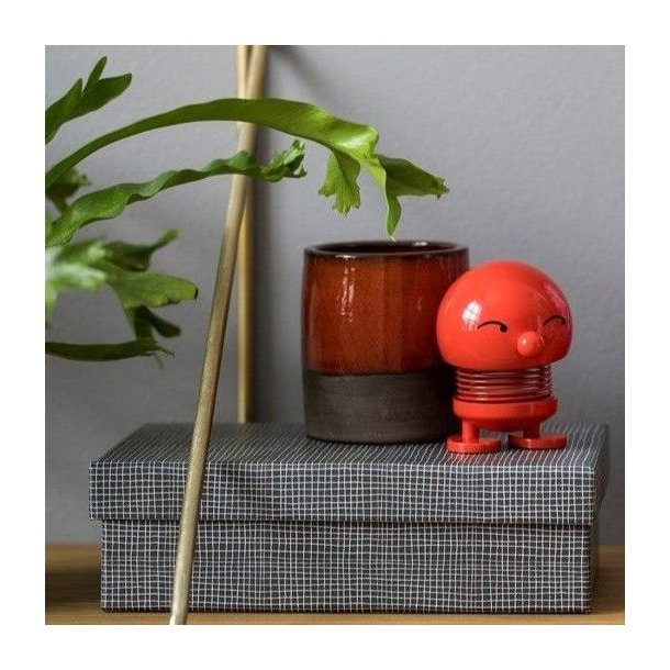hoptimist-bimble-large-roed-plastfigur-miljoe En klassisk r�d Hoptimist Bimble pige i plast med en h�jde p� 15 cm og en diameter p� 10 cm st�ende dekorativt i et milj� p� en m�nstret �ske ved siden af en brun urtepotte med en gr�n plante foran en gr� v�g.