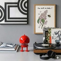 hoptimist-bumble-large-roed-pladespiller-hygge-miljoe Firkantet stemningsbillede af en hylde. Her ses en stor, r�d Bumble Hoptimist med begge �jne �bne, der st�r ved siden af en vintage pladespiller med hovedtelefoner og flere vinylplader. I baggrunden ses en stor sort/hvid grafisk plakat og en mindre plakat med en bee og syrener, der understreger den hyggelige og retro-inspirerede indretning.