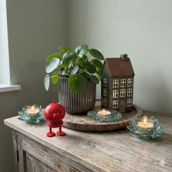 En samling af fire unikke gr�nlige glasfyrfadsstager, en m�rk ribbed keramikurtepotte med en frodig Pilea-plante, og et m�rkegr�nt keramikhus arrangeret p� en rustik tr�overflade, med en lille r�d Hoptimist figur i forgrunden, i et kvadratisk milj�billede.