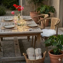 En hyggelig krog p� en terrasse med et d�kket bord og en naturfarvet fletstol, hvor en hvid firkantet lammeskindshynde er lagt p� s�det for at skabe en bl�d og indbydende siddeplads.