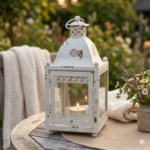 En rustik hvid metal lanterne med patina og et t�ndt fyrfadslys, placeret p� et tr�bord omgivet af hyggelige plaider og en buket med lavendel i en have.