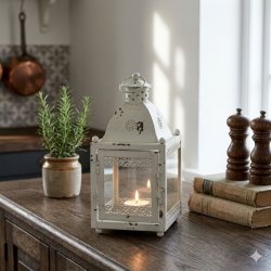 Et kvadratisk 600x600 stemningsbillede, der viser den patinerede hvide metal lanterne fra image_29.png, her placeret p� en m�rk eg k�kkensk�nk. Lanternen st�r centralt og indeholder et t�ndt fyrfadslys, der kaster en flakkende, varm gl�d og lyser op fra glasets base. Surrounding lanternen p� sk�nkens tr�overflade er nogle omhyggeligt udvalgte, naturlige k�kkenrelaterede elementer: en krukke med friske krydderurter og en lille stak gamle kogeb�ger. Baggrunden er sl�ret med en tapet af m�nstret fliser, der understreger en rustik og hyggelig k�kkenatmosf�re. Lysset er varmt og bl�dt, og fremh�ver metallets patina