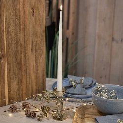 H�jt, slankt hvidt LED-stagelys med t�ndt, varm flamme, placeret i en glasholder p� et d�kket bord med tr�detaljer og en bl� sk�l.
