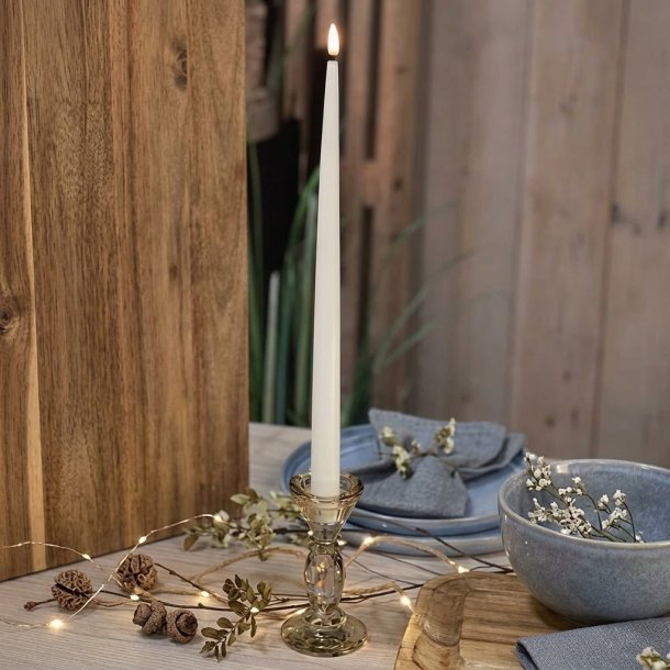 H�jt, slankt hvidt LED-stagelys med t�ndt, varm flamme, placeret i en glasholder p� et d�kket bord med tr�detaljer og en bl� sk�l.