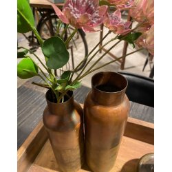 Et s�t med to kobberfarvede jernvaser i forskellige h�jder, den ene med en gren af kunstige, rosa blomster i, placeret p� en hvid kaminhylde med en rustik b�nk i baggrunden.