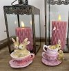 To forskellige p�skefigurer med kaniner i lyser�de kopper opstillet sammen med rosa bloklys i rustikke lanterner.