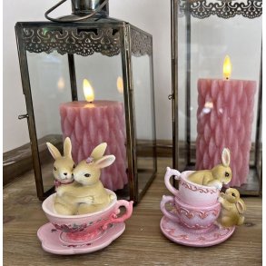 To forskellige p�skefigurer med kaniner i lyser�de kopper opstillet sammen med rosa bloklys i rustikke lanterner.