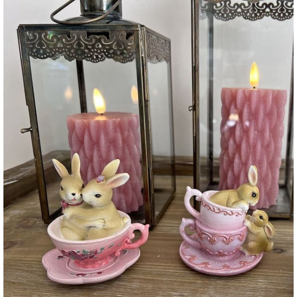 To forskellige p�skefigurer med kaniner i lyser�de kopper opstillet sammen med rosa bloklys i rustikke lanterner.