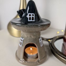 N�rbillede af et keramikhus med m�rkt spidst tag og beige base brugt som fyrfadsstage. Et t�ndt fyrfadslys ses gennem d�r�bningen og skaber et varmt lys i huset, som er placeret i en hyggelig opstilling med gulddetaljer i baggrunden.