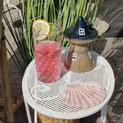 Stemningsbillede med et For�rs Skovhus i beige og m�rk keramik p� et hvidt havebord. Ved siden af huset st�r et glas med lyser�d drink og citronskive samt en lyser�d muslingeskal med h�jt gr�s i baggrunden.