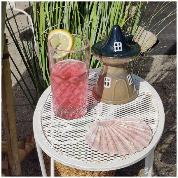 Stemningsbillede med et For�rs Skovhus i beige og m�rk keramik p� et hvidt havebord. Ved siden af huset st�r et glas med lyser�d drink og citronskive samt en lyser�d muslingeskal med h�jt gr�s i baggrunden.