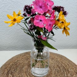 Klar rillet glasvase fyldt med en farverig buket af lyser�de og gule sommerblomster placeret p� en flettet d�kkeserviet.