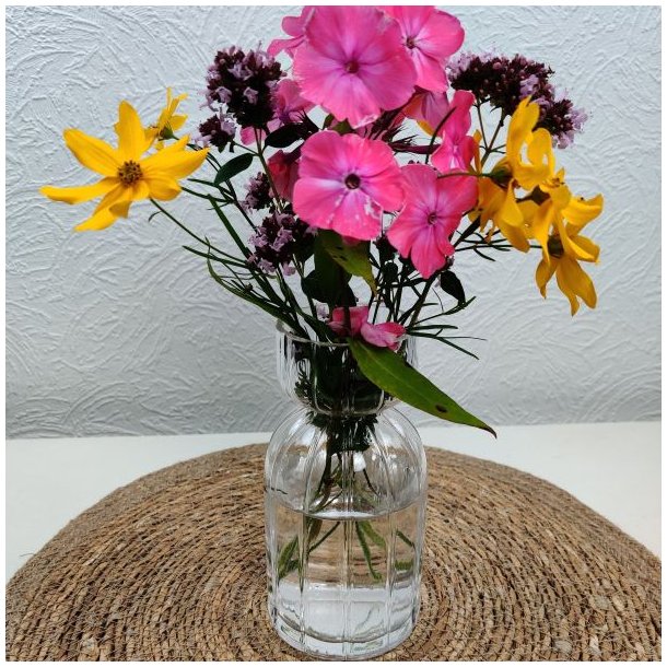 Klar rillet glasvase fyldt med en farverig buket af lyser�de og gule sommerblomster placeret p� en flettet d�kkeserviet.