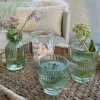Gr�nne La Vida fyrfadsstager og vaser i rillet glas opstillet p� en flettet bakke udend�rs med blomster.