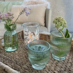 Gr�nne La Vida fyrfadsstager og vaser i rillet glas opstillet p� en flettet bakke udend�rs med blomster.
