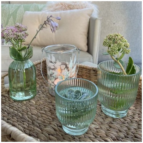 Gr�nne La Vida fyrfadsstager og vaser i rillet glas opstillet p� en flettet bakke udend�rs med blomster.