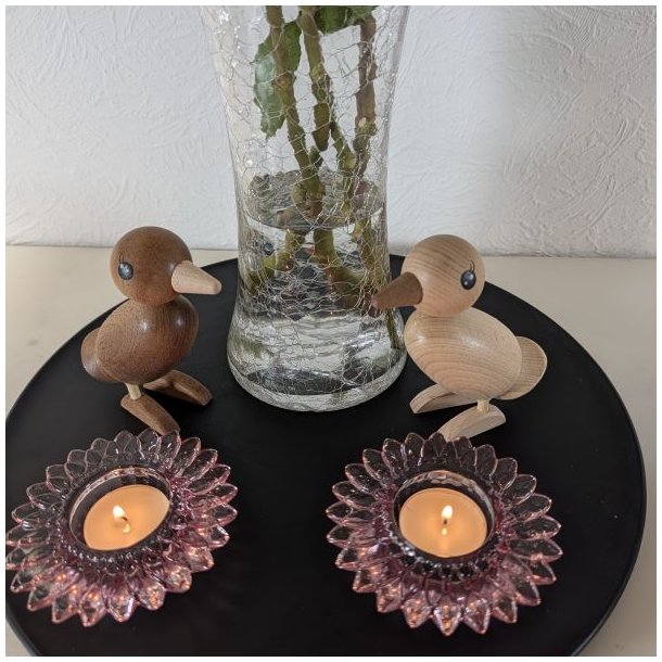 En skarp close-up-fotografering af et par tr�fugle-figurere, der minder om B�lling-�nder. Den m�rkere, r�gede egetr�sfugl til venstre og den lysere egetr�sfugl til h�jre st�r over for hinanden p� en sort metalbakke og danner et par. Baggrunden er st�rkt sl�ret, men antyder den krakelerede glasvase. Fuglenes tr�tekstur og malede �jne er i fokus under bl�dt, naturligt lys. Billedet er kvadratisk.