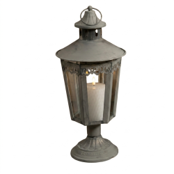 Fritlagt produktbillede af en antik gr� metal-lanterne p� en h�j, dekorativ fod. Lanternen har glaspaneler, en ornamenteret top og et ringh�ndtag. Inden i lanternen st�r et hvidt bloklys, som er t�ndt og afgiver en varm flamme. Hele lanternen er vist mod en ren, hvid baggrund i et kvadratisk 1:1 format.