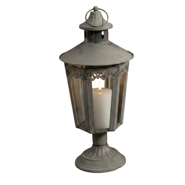 Fritlagt produktbillede af en antik gr� metal-lanterne p� en h�j, dekorativ fod. Lanternen har glaspaneler, en ornamenteret top og et ringh�ndtag. Inden i lanternen st�r et hvidt bloklys, som er t�ndt og afgiver en varm flamme. Hele lanternen er vist mod en ren, hvid baggrund i et kvadratisk 1:1 format.