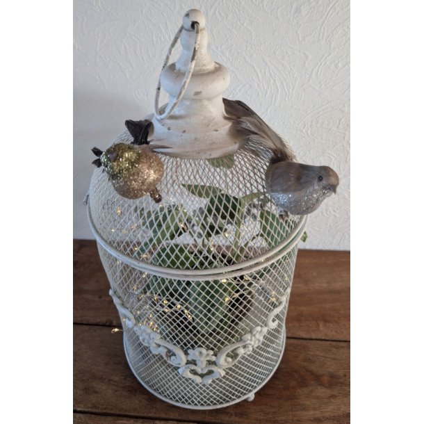 N�rbillede af et hvidt lanternebur i metalnet dekoreret med to sm� glitrende pyntefugle. Indeni buret ses en gr�n plante og en fin lysk�de, der skaber en hyggelig atmosf�re.