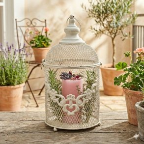 Det antikke, hvide metal-lanternebur (fra image_95.png-image_97.png) st�r smukt dekoreret p� et vejrslidt tr�gulv p� en solrig altan. Indeni buret er en lyser�d lys-cylinder (fra image_96.png) toppet med en fin, t�rret dekoration af lavendel, rosenblade og kviste, omgivet af friske kviste af timian og rosmarin. I baggrunden ses en Mediterranean-style altan med terracotta-krukker og blomstrende planter i det bl�de dagslys. Billedet er kvadratisk (1:1).