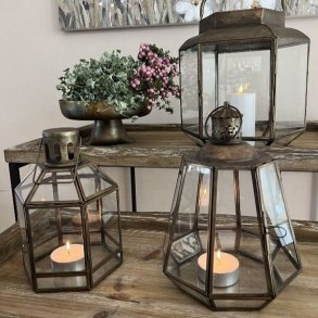 To geometriske, antikbrune metal- og glaslanterner med t�ndte fyrfadslys opstillet p� et rustikt tr�bord i en hyggelig stue.