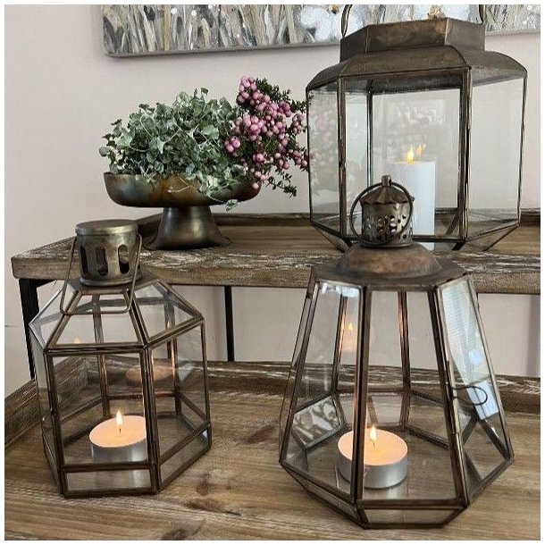 To geometriske, antikbrune metal- og glaslanterner med t�ndte fyrfadslys opstillet p� et rustikt tr�bord i en hyggelig stue.