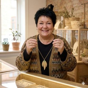 En smilende, �ldre kvinde med kort sort h�r st�r i en smykkeforretning og viser den forgyldte La Vida guldhalsk�de med det trebladede tr�dnet-vedh�ng og den gr�nne stenperle frem.