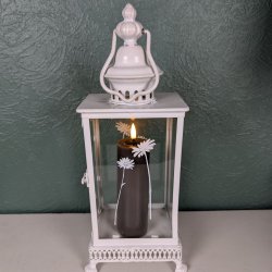 Stemningsfuldt milj�billede af det mokkabrune LED-bloklys med t�ndt real flamme fra Deluxe Homeart, placeret indeni en hvid metal- og glaslanterne dekoreret med hvide blomstermotiver.