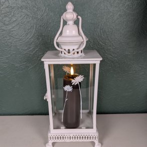 Stemningsfuldt milj�billede af det mokkabrune LED-bloklys med t�ndt real flamme fra Deluxe Homeart, placeret indeni en hvid metal- og glaslanterne dekoreret med hvide blomstermotiver.