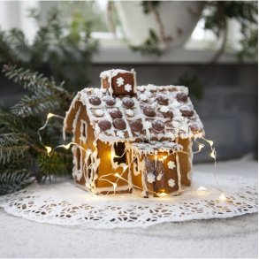 En hyggelig julescene med en fin LED-lysk�de, der lyser op omkring et fint pyntet peberkagehus placeret p� en hvid blonde-serviet.