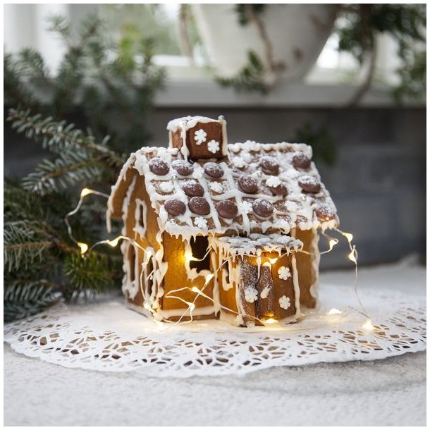 En hyggelig julescene med en fin LED-lysk�de, der lyser op omkring et fint pyntet peberkagehus placeret p� en hvid blonde-serviet.