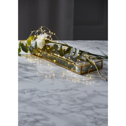 En elegant dekoration p� et marmorbord, hvor en LED-lysk�de ligger i en aflang guldbakke sammen med en hvid blomst og gr�nne blade.
