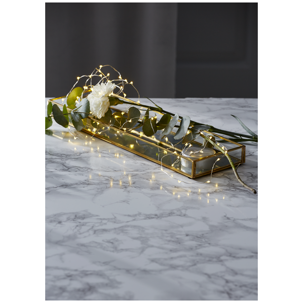 En elegant dekoration p� et marmorbord, hvor en LED-lysk�de ligger i en aflang guldbakke sammen med en hvid blomst og gr�nne blade.