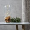 En dekorativ ops�tning, hvor en t�ndt varm-hvid LED-lysk�de er placeret i en h�j glaskrukke fyldt med valn�dder p� et hvidt bord.