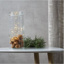 En dekorativ ops�tning, hvor en t�ndt varm-hvid LED-lysk�de er placeret i en h�j glaskrukke fyldt med valn�dder p� et hvidt bord.