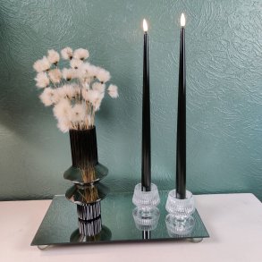 Milj�billede af to t�ndte sorte LED-stagelys i klare glaslysestager st�ende p� en spejlbakke ved siden af en m�rk vase med hvide blomster mod en gr�n v�g.