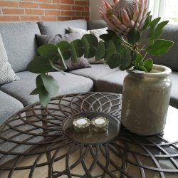 Stemningsbillede fra en dagligstue, hvor to sm�, detaljerede glas fyrfadsstager i antik s�lv med t�ndte lys st�r p� et sofabord ved siden af en stor vase med eucalyptus og en protea-blomst.