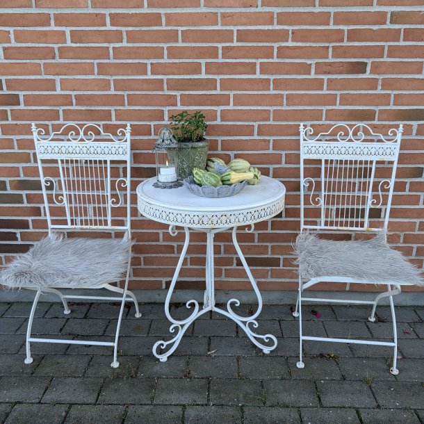 Et s�t af to hvide caf�stole i metal med matchende lysegr� imiterede lammeskindshynder, opstillet omkring et lille hvidt havebord mod en murstensv�g.