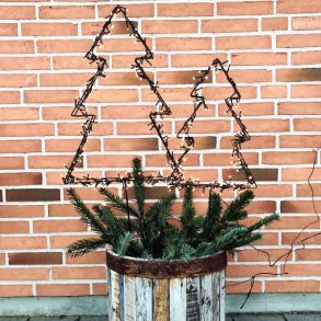 Et kvadratisk milj�billede i 1:1 format, der viser en dekorativ udend�rs juledekoration. To stilistiske juletr�er formet af m�rkt metalknap med p�monterede t�ndte LED-lysk�der er stukket ned i en rustik, patineret tr�-halvt�nde med rustne metalb�nd. Halvt�nden er fyldt med friske grangrene. Dekorationen er placeret mod en klassisk, r�d murstensv�g, og julelysene kaster et varmt og hyggeligt sk�r i skumringen. Man ser murstensv�ggens tekstur, og julelysene st�r tydeligt frem.