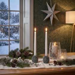Stemningsfuldt firkantet milj�billede af et s�t med to grankogle lysestager med t�ndte hvide stagelys placeret p� et bord foran et vindue og m�nstret tapet.