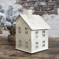 Et stemningsfuldt milj�billede i 1:1 format af et lyshus i antique creme jern placeret p� et rustikt tr�bord. Ved siden af huset st�r et lille sned�kket juletr�, og baggrunden viser en r� murstensv�g, der fremh�ver husets antikke finish.