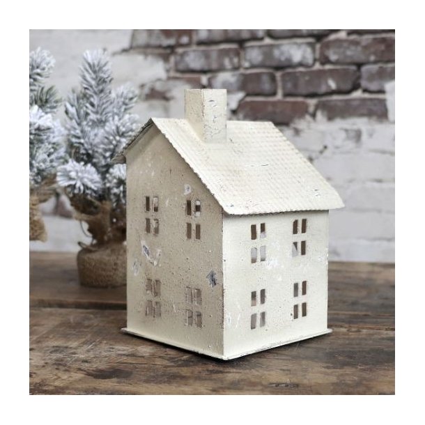 Et stemningsfuldt milj�billede i 1:1 format af et lyshus i antique creme jern placeret p� et rustikt tr�bord. Ved siden af huset st�r et lille sned�kket juletr�, og baggrunden viser en r� murstensv�g, der fremh�ver husets antikke finish.