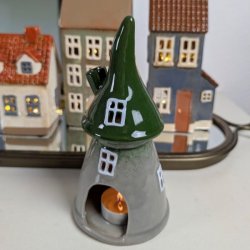 Keramik Skovhus med et t�ndt fyrfadslys indeni, der lyser vinduerne op, vist i en opstilling med andre sm� keramikhuse.