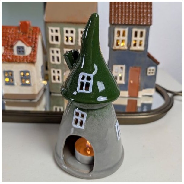 Keramik Skovhus med et t�ndt fyrfadslys indeni, der lyser vinduerne op, vist i en opstilling med andre sm� keramikhuse.