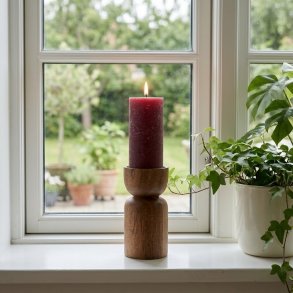 Macon rustikt bordeauxr�dt bloklys H20 cm med t�ndt flamme placeret i en tr�lysestage i en lyst vindueskarm ved siden af en gr�n potteplante.