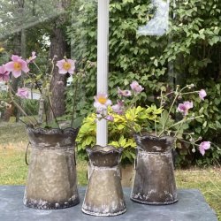 Tre meleret zinkurtepotter med dekorative hanke og b�lget kant, her brugt som vaser til lyser�de h�stanemoner. Potterne er placeret p� et m�rkt bord i en gr�n have under en ranke med m�rke vindruer, hvilket fremh�ver deres rustikke patina.