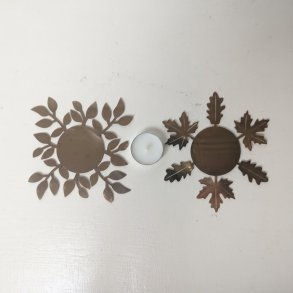 Et top-down produktbillede af to forskellige dekorative, flade fyrfadsstager i m�rk metal, placeret side om side p� en hvid tr�overflade. Den venstre stage er formet med en t�t ring af mindre, ovale blade. Den h�jre stage er formet med tydelige ahornsblade og egetr�sblade. Mellem de to stager st�r et t�ndt hvidt fyrfadslys. Billedet er kvadratisk.
