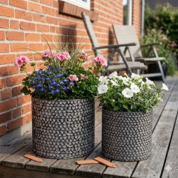 Et stemningsfuldt milj�billede i 1:1 format, der viser de to m�nstrede metalkrukker fra image_98.png og image_99.png, placeret sammen som en gruppe p� et tr�gulv p� en terrasse. Den store krukke (H26,5 cm) og den lille krukke (H23,5 cm) er beplantet med blomstrende pelargonier og petunier, samt dekorativt prydgr�s. Baggrunden viser en r�d murstensv�g og sl�ret havevegetation i sollys.